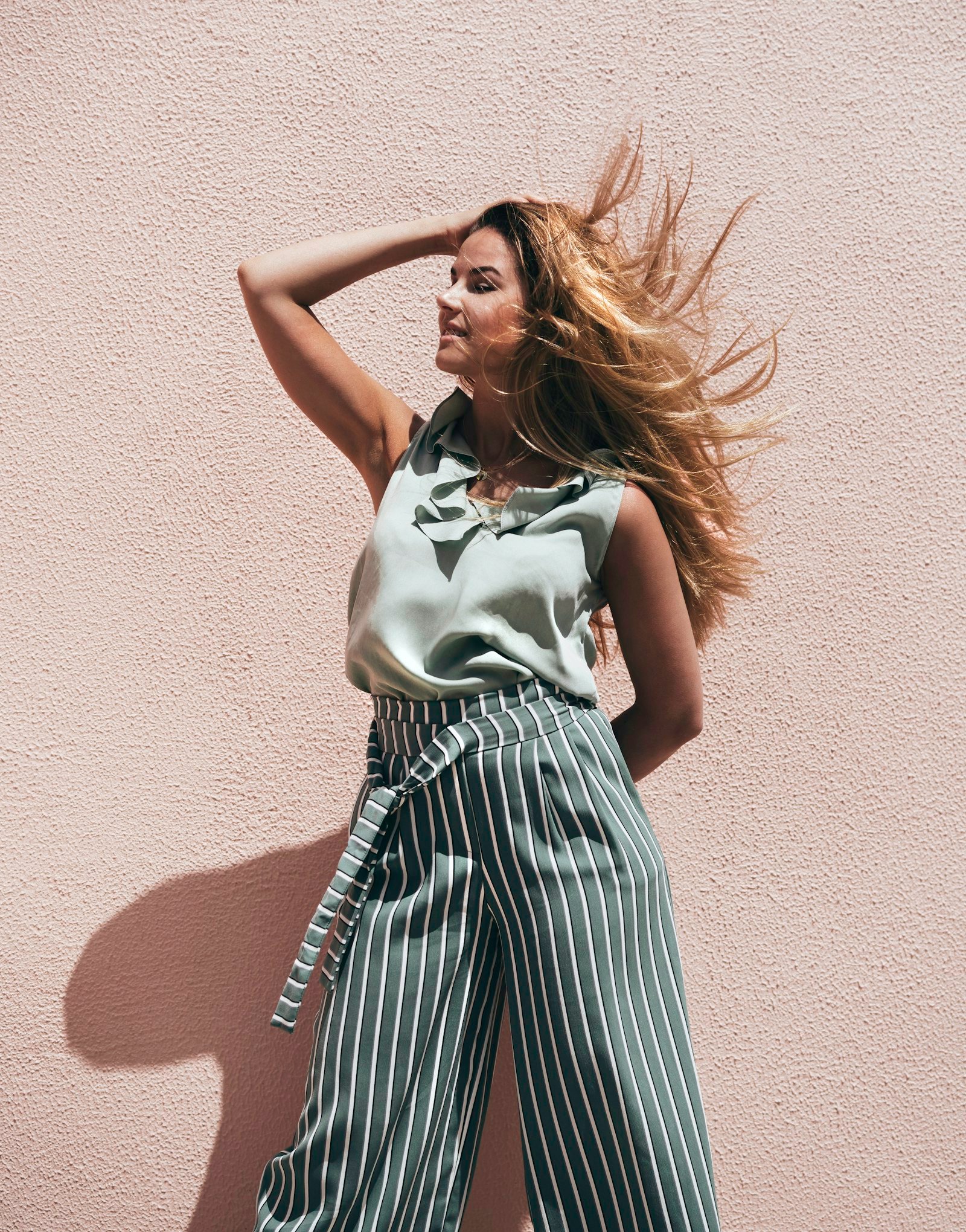 Mulher com cabelo ao vento, blusa verde-sálvia e calça pantalona listrada, diante de uma parede rosa ao sol